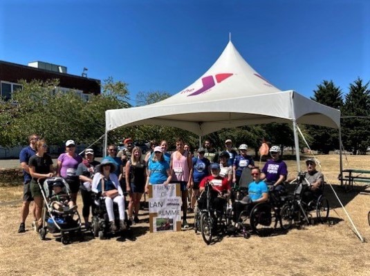 A crowd of approximately 20 people under an event tent, some in wheelchairs, encircling a sign that reads 'Friends of Lan Remme'